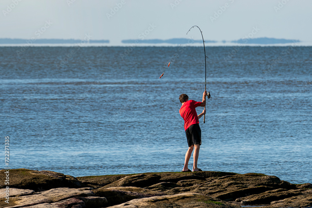 Young man standing on the jetty casting fishing line into the ocean on sunny summer day