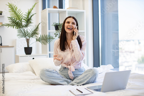 Caucasian brunette using modern devices enjoying break from work for business negotiations on weekend. Beautiful woman relaxing on bed at home and using modern smartphone for talking with family.