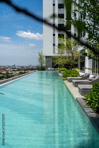 A serene rooftop swimming pool extends towards the horizon, bordered by modern lounge chairs and vibrant greenery, against a backdrop of cityscape and clear blue skies..