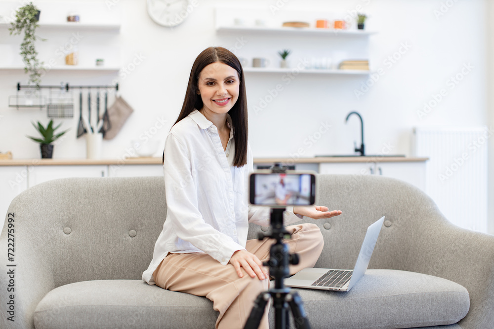 Front view of cheerful content maker in casual wear recording video to followers on modern smartphone. Young caucasian woman in with laptop sitting on comfortable sofa in kitchen. Front view of cheerful content maker in casual wear recording video to followers on modern smartphone. Young caucasian woman in with laptop sitting on comfortable sofa in kitchen.