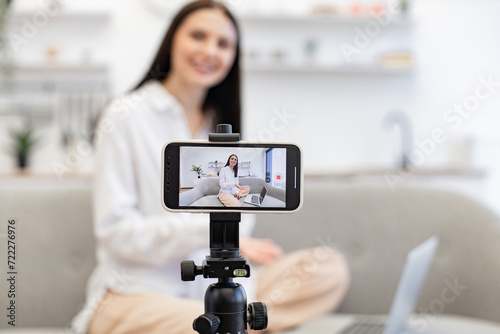 Positive caucasian female influencer recording video on modern smartphone while sitting on couch with laptop. Happy young woman sharing her success with subscribers online. © sofiko14