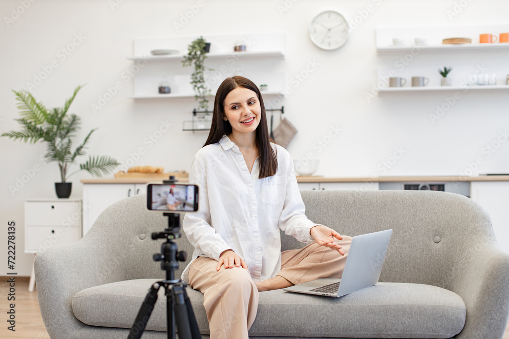 Caucasian woman with brunette hair reading information from modern laptop during live broadcast on social network. Demanding tutor using modern smartphone with tripod for online training. Caucasian woman with brunette hair reading information from modern laptop during live broadcast on social network. Demanding tutor using modern smartphone with tripod for online training.