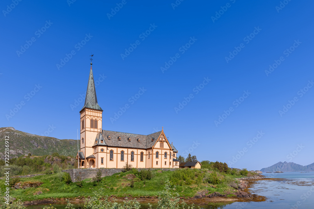 Vagan Kirke, a striking historical church in Lofoten, Norway, stands proudly by the waterside, its spire reaching into the clear sky amid a serene natural setting
