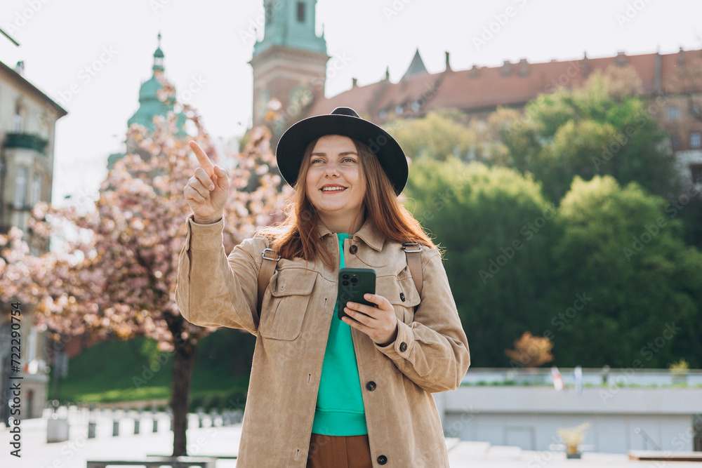 Attractive young female tourist in hat is exploring new city. Redhead 30s girl with smartphone pointing finger near Wawel castle, Cracow, Poland. Traveling Europe in spring