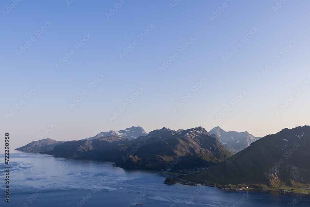 Evening descends on the Lofoten Islands, Norway, with the setting sun casting a warm glow on the imposing mountain range and the tranquil Norwegian Sea hugging the coastline
