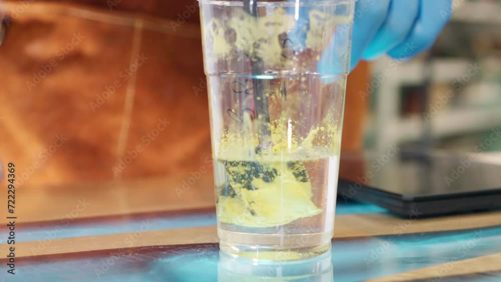 Trendy transparent epoxy resin in plastic cup mixing with yellow colour pigment to create multi-component solution for coating individual wooden tabletops. Industrial mixer stirring two substances to Trendy transparent epoxy resin in plastic cup mixing with yellow colour pigment to create multi-component solution for coating individual wooden tabletops. Industrial mixer stirring two substances to