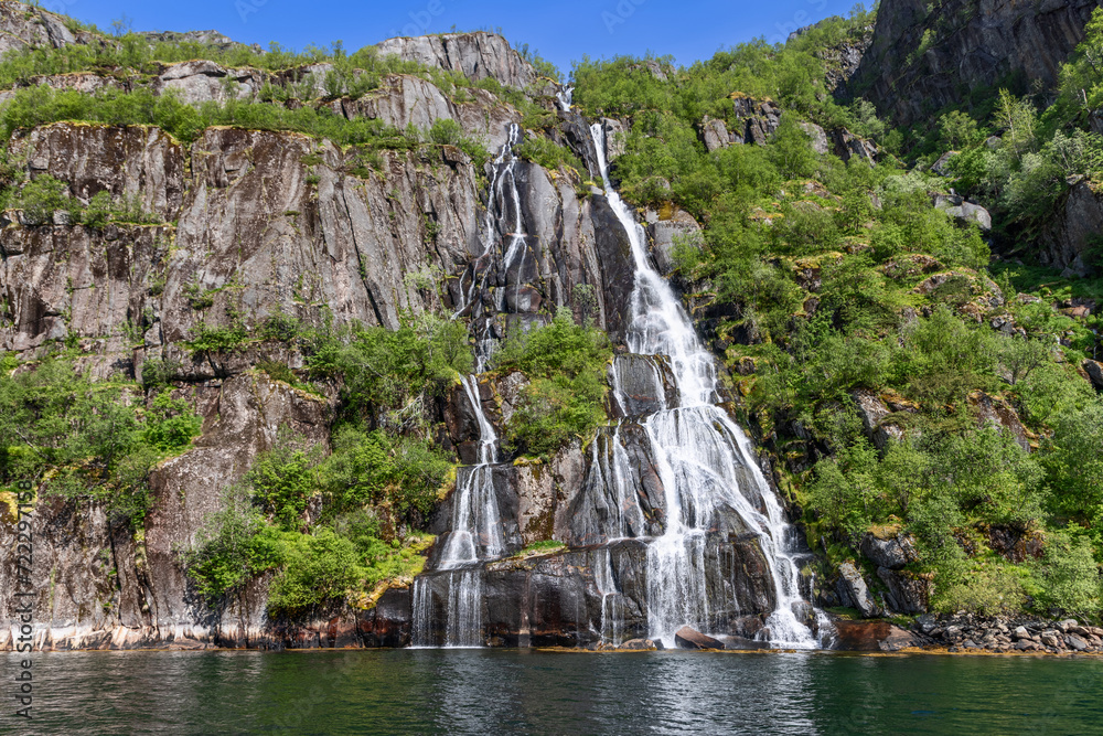 Fototapeta premium A striking waterfall splits into silvery threads down a moss-draped cliff into the emerald waters of Trollfjorden in Lofoten, Norway