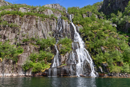 A striking waterfall splits into silvery threads down a moss-draped cliff into the emerald waters of Trollfjorden in Lofoten, Norway