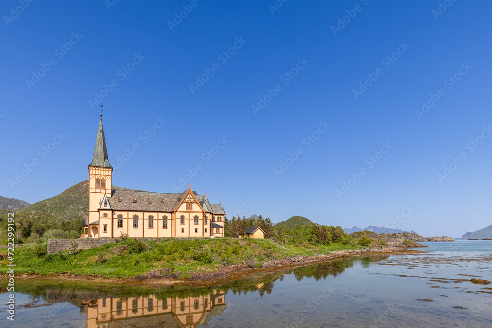 Vagan Kirke, nestled in Lofoten's stunning landscape, reflects its traditional architecture in the tranquil waters on a bright Norwegian day