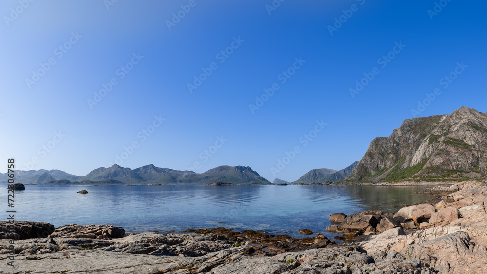 Lofoten's rugged coastline unfolds under a clear sky, with mountains rising behind the serene Norwegian Sea, a pristine example of Norway's natural splendor