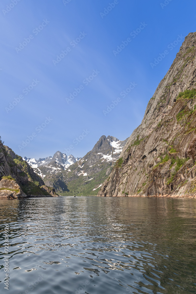 A vertical view of the serene Trollfjorden in Lofoten, Norway, where steep cliffs meet the calm waters, with snow patches adorning the mountain peaks