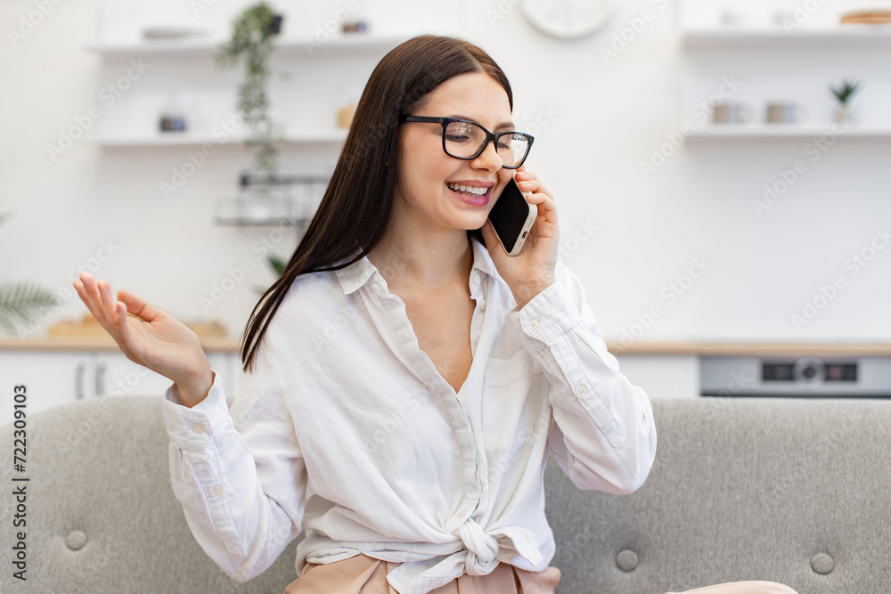 Charming woman in casual attire enjoying communication with colleagues while sitting on cozy sofa. Positive caucasian female gesturing while having conversation on modern smartphone, staying at home. Charming woman in casual attire enjoying communication with colleagues while sitting on cozy sofa. Positive caucasian female gesturing while having conversation on modern smartphone, staying at home.