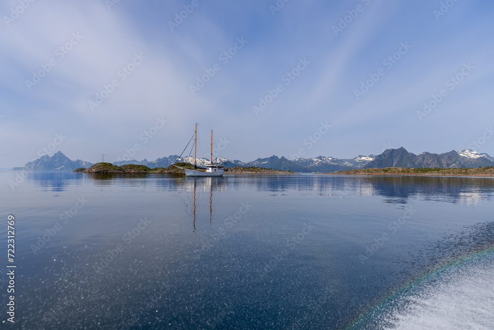 Calm sea reflects a sailboat and rugged Norwegian mountains, with a subtle rainbow arc from water splashes in the tranquil Lofoten region