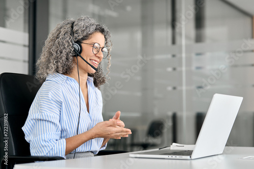 Smiling middle aged female support service employee talking to customer sitting at desk. Happy senior old woman professional call contact center agent wearing headset hybrid working in business office photo