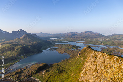 The sun casts a soft glow over the serene lakes and rugged hills of Vestvagoya, seen from the rocky crest of Offersoykammen in the Lofoten archipelago