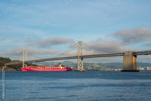 a large red container ship making its way under the Oakland Bay Bridge in daylight on a partly cloudy day. © kmlPhoto