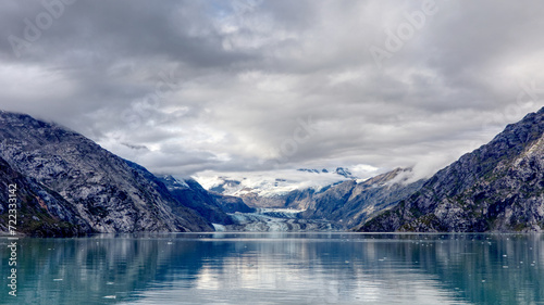 Johns Hopkins Glacier in Glacier Bay Alaska. Majestic mountains and clouds frame the glacier with reflections in the foreground. 