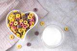 sweet multi-colored cereal rings for a healthy breakfast in a heart-shaped plate, milk in a transparent double-walled mug.Top view