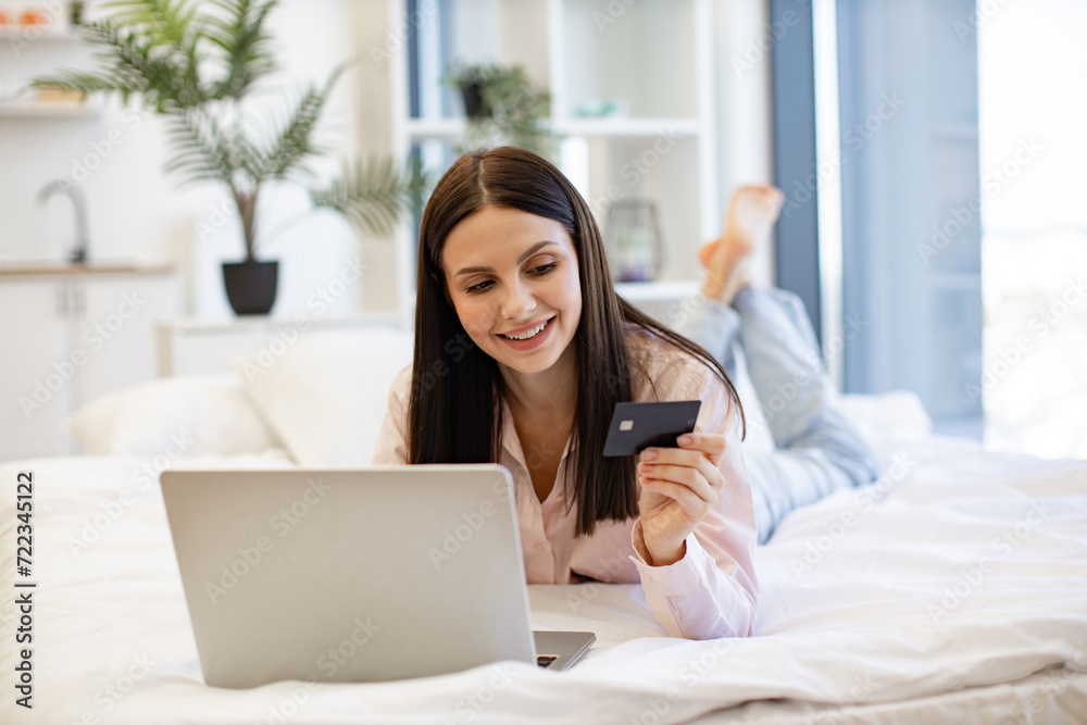 Relaxed lady lying on comfy bed and enjoying purchase of goods on Internet using bank card at home on weekend. Smiling young woman making online order using modern wireless laptop. Relaxed lady lying on comfy bed and enjoying purchase of goods on Internet using bank card at home on weekend. Smiling young woman making online order using modern wireless laptop.