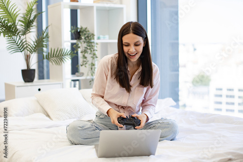 Beautiful young woman relaxing on cozy bed at home and using modern wireless joystick for playing games. Caucasian brunette using modern devices enjoying favorite video game on weekend.