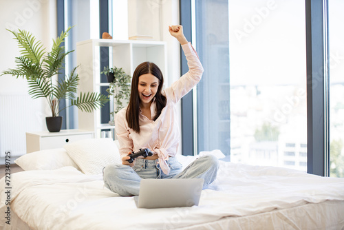 Young woman raising hands with wireless joystick while win in video games using modern laptop. Happy lady sitting on comfy bed and celebrating victory in favorite computer game on weekend.