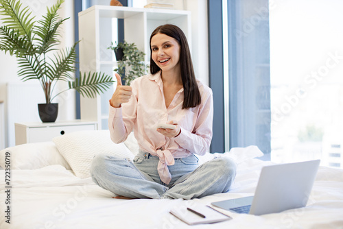 Attractive young woman working remotely from home, typing on modern wireless cell phone, thumb up. Relaxed lady sitting on comfy bed and enjoying chatting with coworkers using smartphone on weekend.