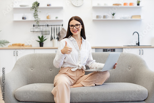 Happy brunette businesswoman sitting on comfy couch and using portable laptop showing thumb up. Excited caucasian female feeling inspired with new idea during freelancing at cozy home. Free Download Happy brunette businesswoman sitting on comfy couch and using portable laptop showing thumb up. Excited caucasian female feeling inspired with new idea during freelancing at cozy home. - GFXLoad