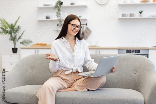 Free Download Charming female freelancer sitting on couch and using modern laptop for video conference at home. Confident brunette woman in talking on webcam and gesturing with hands. - GFXLoad