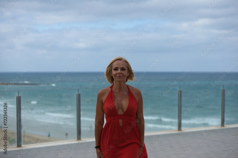 Beautiful blonde adult woman in an elegant red dress walks along the viewpoint above the beach. In the background the Atlantic Ocean in Cadiz, Spain. Travel and holiday concept. Beautiful blonde adult woman in an elegant red dress walks along the viewpoint above the beach. In the background the Atlantic Ocean in Cadiz, Spain. Travel and holiday concept.