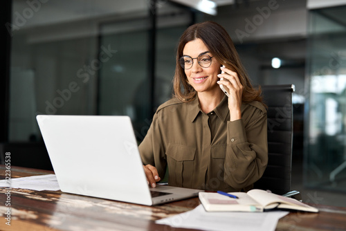 Happy smiling mature middle aged business woman, busy professional hr or bank manager talking on the phone making business call on cellphone at work in office using laptop computer.