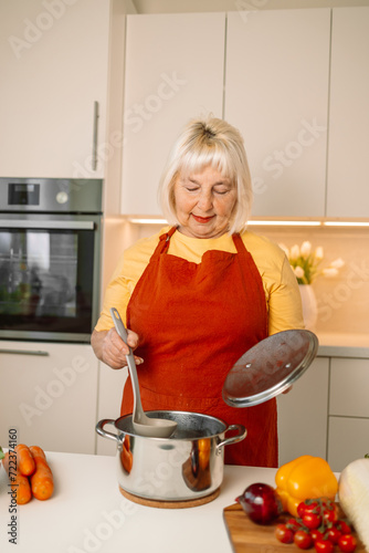 Beautiful happy senior woman is cooking in the home kitchen and testing some soup from the pan on the stove. High quality photo