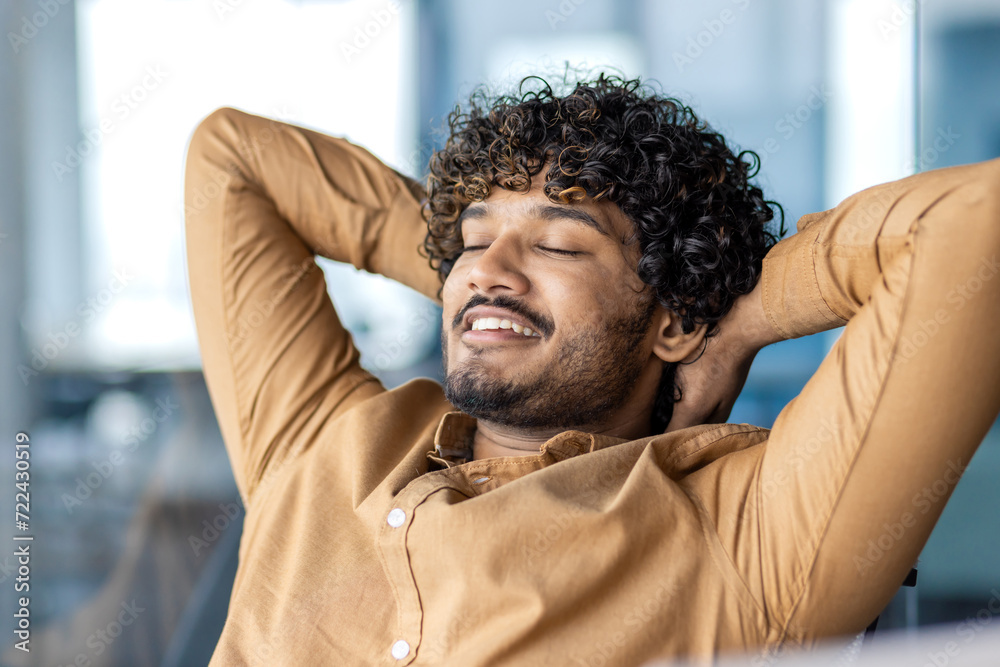 Portrait of relaxed latino guy in camel shirt leaning on chair back and holding hands behind head with wide smile. Productive entrepreneur having break from working on startup with impressive profit. - obrazy, fototapety, plakaty 