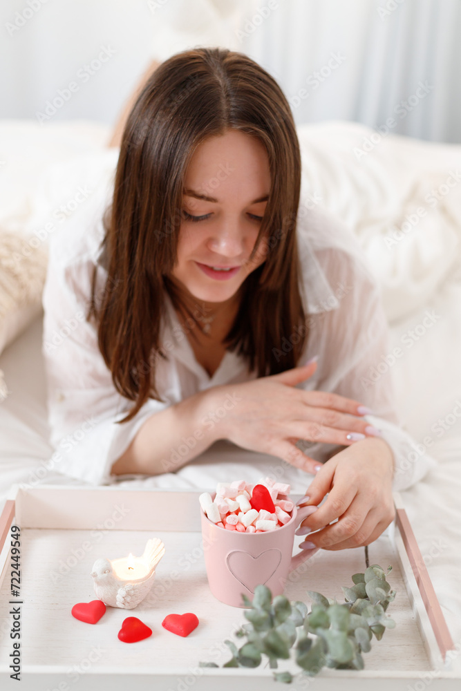 Beautiful woman with pink mug of cocoa, marshmallows and heart-shaped candy on bed at home. St Valentine's Day. Beautiful woman with pink mug of cocoa, marshmallows and heart-shaped candy on bed at home. St Valentine's Day.