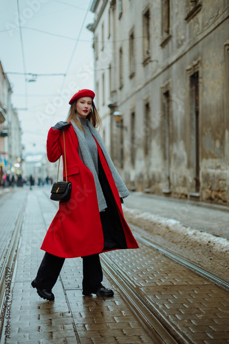 Fashionable confident woman wearing elegant red midi long coat, beret, gray knitted scarf, carrying trendy black leather purse, walking in street of European city. Outdoor full-length fashion portrait