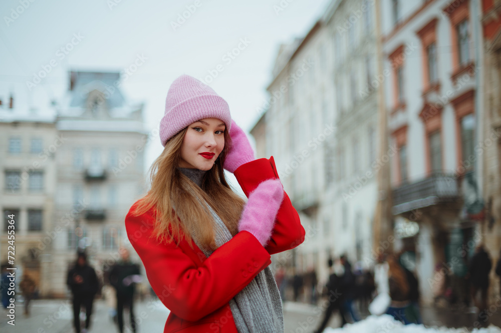Fashionable happy smiling woman wearing pink beanie hat, mittens, red coat, gray scarf, posing in street of European city. Christmas, outdoor winter holidays concept. Copy, empty, blank space for text
