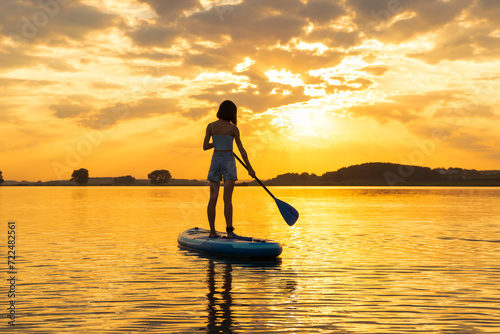 silhouette of woman on inflatable SUP board and paddling through shining water surface. aesthetically wide shot. Freedom happy female at sunset on a lake © Yekatseryna