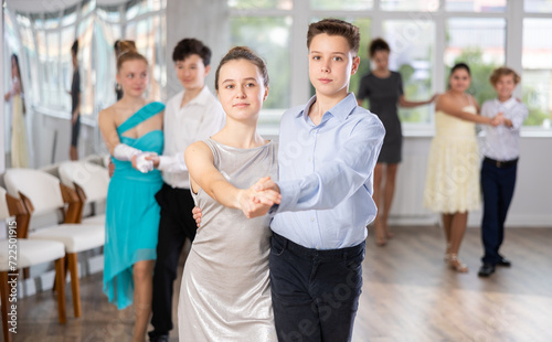 Emotional adolescent ballroom dancers, girls and boys in formal wear practicing elegant dance moves in pairs in bright studio, with female instructor supervising in background..