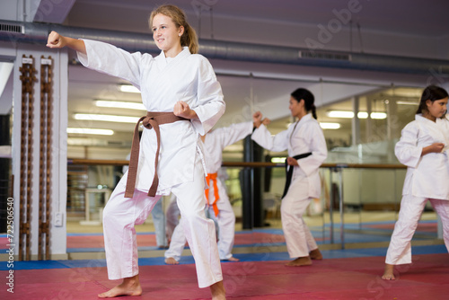 Sporty girl in kimono posing in gym while other kids sparring in background during karate class