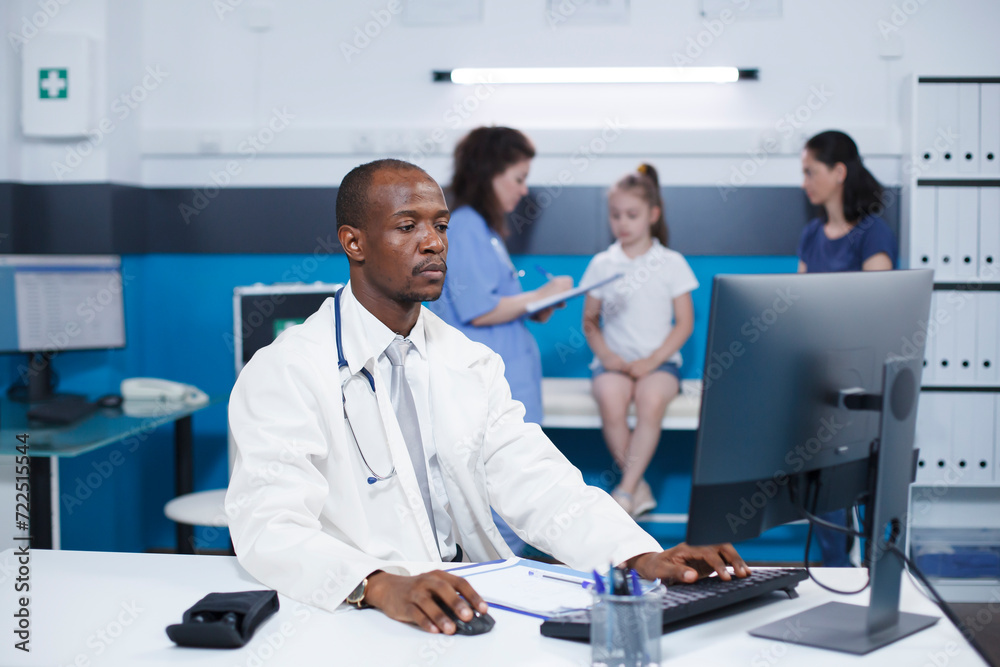 Dedicated African American male doctor using the desktop pc to document his medical notes. In the background, a caucasian nurse attends to a child in a well-equipped examination room. Dedicated African American male doctor using the desktop pc to document his medical notes. In the background, a caucasian nurse attends to a child in a well-equipped examination room.
