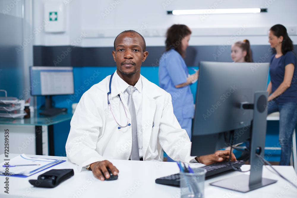 African american male doctor sitting at a desk in hospital office, looking at camera. Portrait shot of black man wearing a lab coat in a clinic while a patient is assisted by nurse in the background. African american male doctor sitting at a desk in hospital office, looking at camera. Portrait shot of black man wearing a lab coat in a clinic while a patient is assisted by nurse in the background.