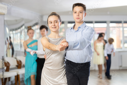 Enthusiastic friendly teenagers, boy and girl in party attire performing elegant waltz in pair in sunny hall of dance school during group lesson..