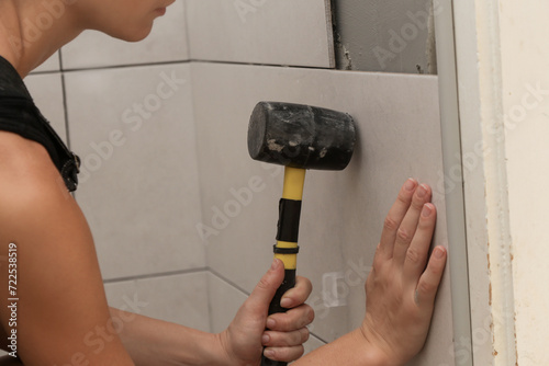 Woman tiling bathroom walls, pressing tile into a glue with rubber hammer. Process of installation of tiles in the bathroom step by step. DIY home improvement. 
