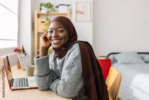 Portrait of young african creative woman smiling at camera sitting at desk. Happy black female student in muslim headscarf relaxing at home. Youth and technology concept. photo