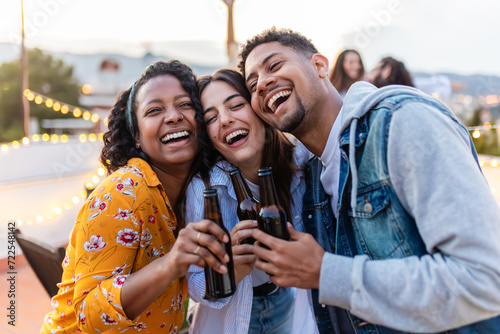 Close-up frontal image of three multi-ethnic young adult friends embracing and drinking beer in an outdoor terrace while celebrating a party. Celebrating together and diversity photo