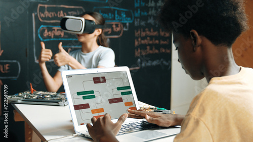 Caucasian school girl wear VR glass and showing thumb up while african boy working on laptop at blackboard coding engineering prompt written in STEM class. Creative education concept. Edification. photo