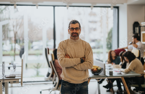 A mature, smiling man with crossed arms stands confidently in an office setup with colleagues interacting in the background.