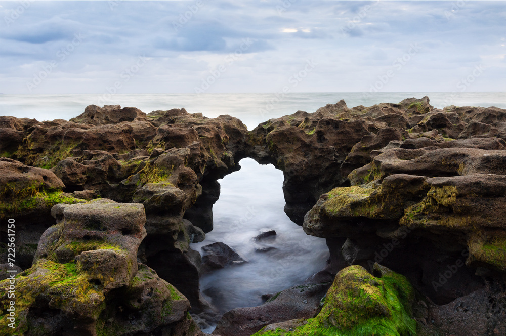 Moss-covered rocks with natural arch on shore, ideal for geology content, eco-tourism marketing, and nature-focused educational material. Moss-covered rocks with natural arch on shore, ideal for geology content, eco-tourism marketing, and nature-focused educational material.
