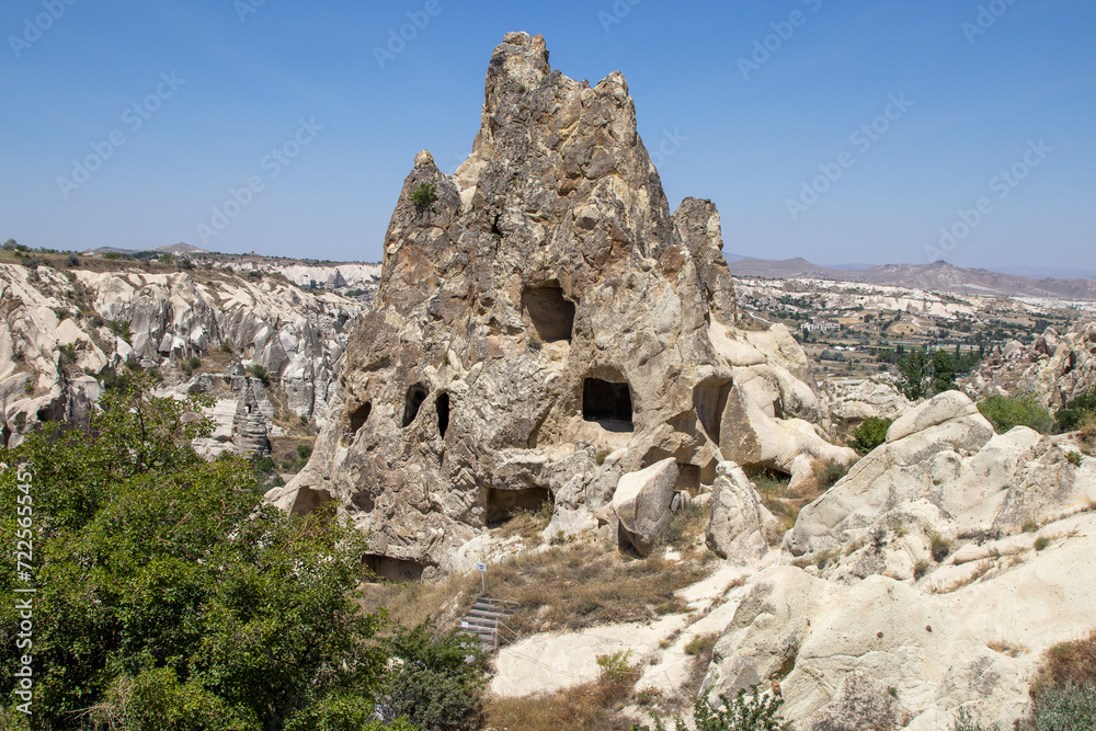 Sunny landscape of Cappadocia’s unique fairy chimneys with cavities, surrounded by green shrubs, under a clear blue sky, highlighting nature’s artistry.