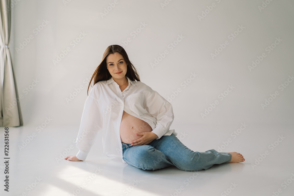 Cute young Pregnant girl sitting on the white studio in warm light with hands on pregnant stomach on wall background with shadow. Pregnant concept, free space, enjoying pregnancy. Pregnant body. Cute young Pregnant girl sitting on the white studio in warm light with hands on pregnant stomach on wall background with shadow. Pregnant concept, free space, enjoying pregnancy. Pregnant body.