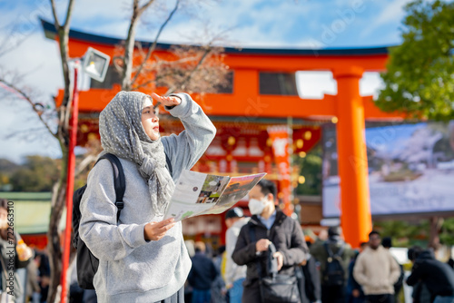 Travel, muslim, Two Asian female tourists of different religions friends visitor learning about history of fushimi inari shrine in travel book while walking through senbon torii path in Kyoto Japan.